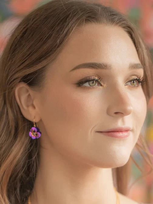 A close-up shows a woman with long brown hair wearing Holly Yashis Sparkling Fuchsia Garden Pansy Drop Earrings. With subtle makeup, she looks slightly to the side as colorful patterns blur softly in the background.