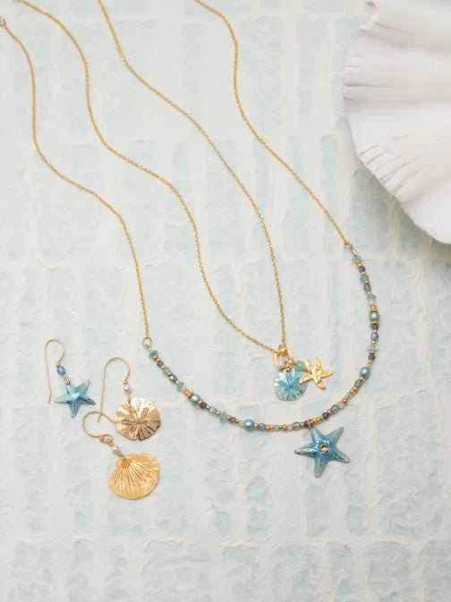 The Holly Yashi Carmel Beaded Seashore Blue Starfish Necklace, displaying two gold star-shaped pendants with blue beads, lies on a textured surface reminiscent of the ocean. Nearby are gold earrings with star and disc designs and a partially visible seashell adding coastal charm.