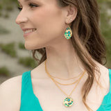 A person with long brown hair and a turquoise top smiles, looking to the side. They wear Seashore Dreams Teal Earrings by Holly Yashi and a pendant necklace, both featuring Austrian crystals in green and gold details, against a blurred green background.