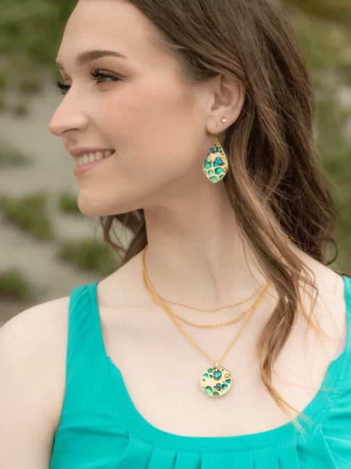 A person with long brown hair and a turquoise top smiles, looking to the side. They wear Seashore Dreams Teal Earrings by Holly Yashi and a pendant necklace, both featuring Austrian crystals in green and gold details, against a blurred green background.