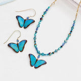 The Ultra Violet Bella Butterfly Beaded Necklace by Holly Yashi features blue butterfly pendants and is adorned with small blue and silver beads. It pairs perfectly with matching earrings, all elegantly presented on a white surface.