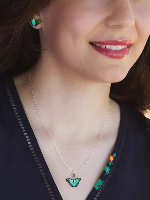 A person wears a smile and the Holly Yashi Green Flash Bella Butterfly Necklace, along with matching earrings. Both feature green butterfly pendants. The necklaces sterling silver chain adds elegance, complementing their subtly patterned dark top as part of their face is visible.