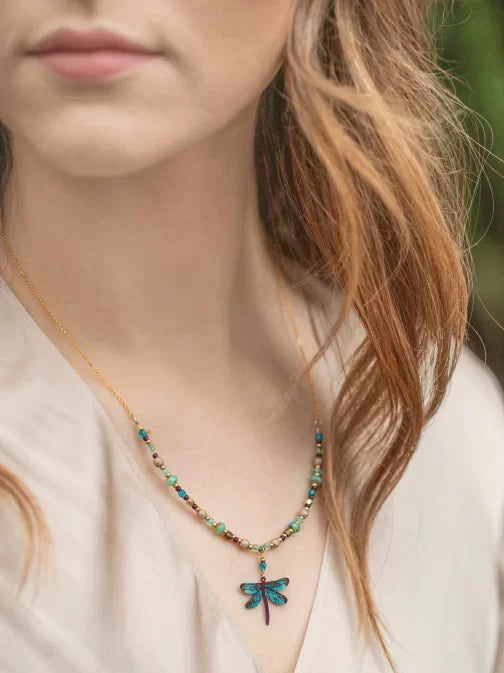 A person with long, wavy hair wears a Turquoise Beaded Dragonfly Dreams Necklace by Holly Yashi. The hypoallergenic piece complements the light blouse, set against a softly blurred background.