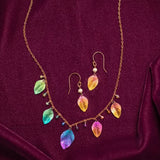 Explore the Healing Leaf Peach Earrings by Holly Yashi Jewelry, showcasing vibrant metallic leaf pendants in hues of blue, green, pink, and gold on a rich burgundy fabric. Elegantly crafted with hypoallergenic niobium chains and delicate bead accents for a sophisticated touch.