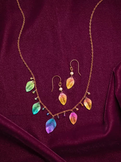 Explore the Healing Leaf Peach Earrings by Holly Yashi Jewelry, showcasing vibrant metallic leaf pendants in hues of blue, green, pink, and gold on a rich burgundy fabric. Elegantly crafted with hypoallergenic niobium chains and delicate bead accents for a sophisticated touch.