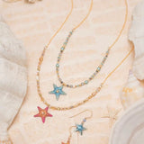The Carmel Beaded Peach Starfish Necklace by Holly Yashi, featuring mother of pearl with gold and blue beads, is displayed on a sandy surface with seashells and coral, evoking a beachy ocean-inspired aesthetic.