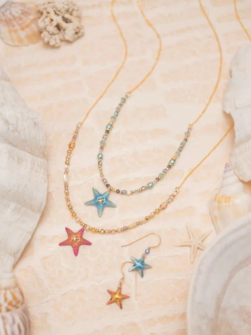 The Carmel Beaded Peach Starfish Necklace by Holly Yashi, featuring mother of pearl with gold and blue beads, is displayed on a sandy surface with seashells and coral, evoking a beachy ocean-inspired aesthetic.