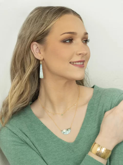 A woman with long, wavy blonde hair smiles while looking to the side. She is wearing a green V-neck sweater and matching hypo-allergenic jewelry with blue accents from Holly Yashi, including Turquoise Tula Earrings and a gold textured bracelet. The background is plain and light-colored.