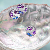 The Ocean Depths Sea Plum Necklace from Holly Yashi features silver circular jewelry set with blue and purple gemstones, elegantly displayed on a pearl-lined shell against a teal background. Paired with earrings, this set is enhanced by Austrian crystals for added sophistication.