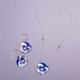 Discover the elegance of this jewelry set featuring a silver chain necklace and Holly Yashis Sparkling Fuchsia Garden Pansy Drop Earrings with stunning purple and white flower pendants on a light purple background.