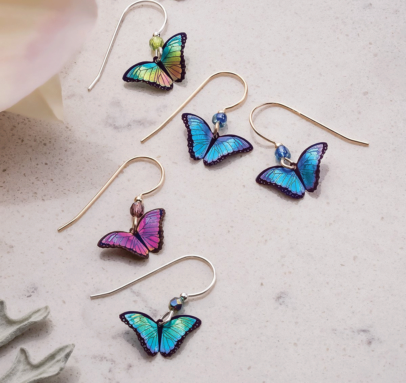 Five pairs of Ultra Violet Butterfly Earrings by Holly Yashi in colors like blue, purple, and green are shown on a light surface. Made from hypo-allergenic niobium, each earring has a decorative bead and thin hook. A softly blurred plant appears partially in the corners.
