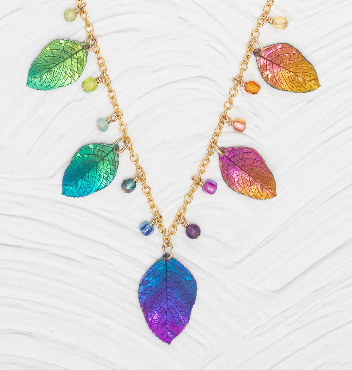 The Healing Elm Leaf Rainbow Necklace by Holly Yashi, 18k gold-plated, showcases iridescent leaf pendants in blue, green, purple, and orange hues with small beads on a textured white base.