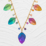 The Healing Elm Leaf Rainbow Necklace by Holly Yashi, 18k gold-plated, showcases iridescent leaf pendants in blue, green, purple, and orange hues with small beads on a textured white base.