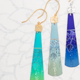 Displayed are three cone-shaped Sage Tula Earrings by Holly Yashi, each with intricate floral designs and gradients: blue to turquoise, silver, and purple. Made with hypo-allergenic niobium metal, they have elegant gold or silver hooks for sensitive ears.