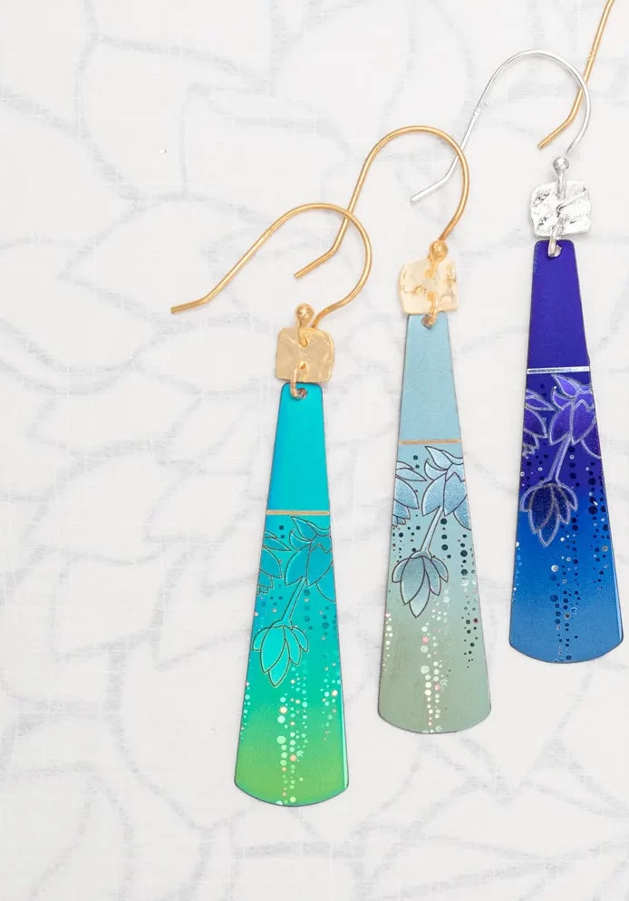 Displayed are three cone-shaped Sage Tula Earrings by Holly Yashi, each with intricate floral designs and gradients: blue to turquoise, silver, and purple. Made with hypo-allergenic niobium metal, they have elegant gold or silver hooks for sensitive ears.