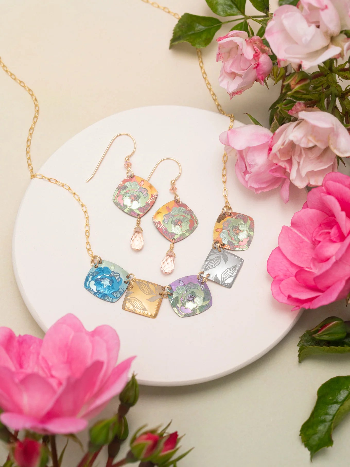The Flora Multi Color Necklace by Holly Yashi, featuring vibrant floral-patterned square pendants, is displayed on a white round plate with pink flowers and green leaves—a stunning handcrafted jewelry set.