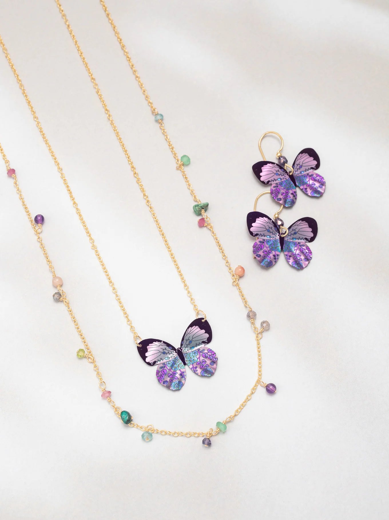 The Holly Yashi Blooming Butterfly Radiant Purple Necklace features an iridescent niobium butterfly pendant with matching earrings and multicolored beads, shown on white. Some pieces offer adjustable gold chains for a perfect fit.