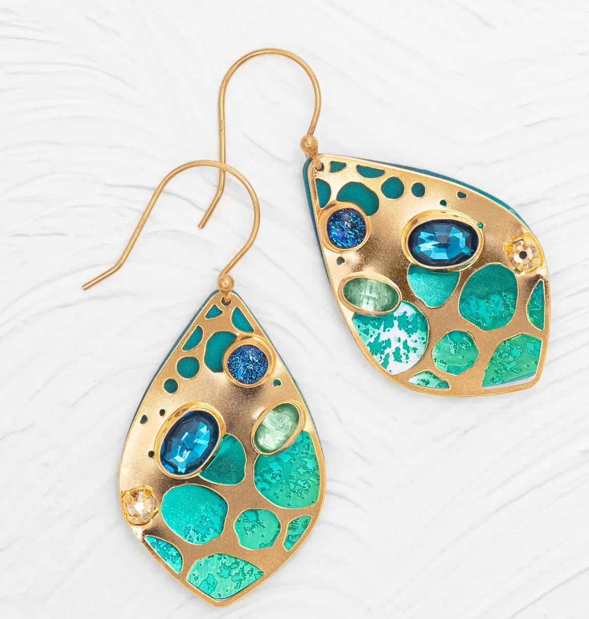Discover the Seashore Dreams Teal Earrings by Holly Yashi - teardrop beauties with gold borders featuring turquoise and blue inlays, adorned with Austrian crystals. The circular design rests elegantly against a textured white background.