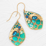Discover the Seashore Dreams Teal Earrings by Holly Yashi - teardrop beauties with gold borders featuring turquoise and blue inlays, adorned with Austrian crystals. The circular design rests elegantly against a textured white background.
