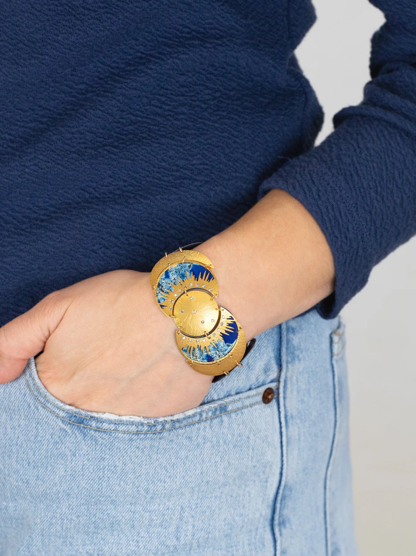 A person in light blue jeans and a navy sweater has their hand in their pocket, highlighting the Holly Yashi Selena Midnight Blue Bracelet—an elegant gold piece with sunburst motifs that brings celestial charm to their wrist.