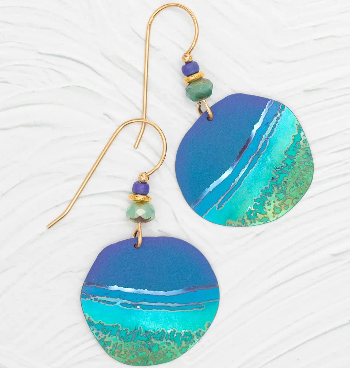 Discover the Rip Tide Calypso Earrings by Holly Yashi, featuring a beach-inspired blue and turquoise wave design. Adorned with green, blue, and gold beads on hypo-allergenic golden hooks, these earrings capture ocean charm against a textured white backdrop.