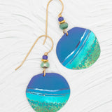 Discover the Rip Tide Calypso Earrings by Holly Yashi, featuring a beach-inspired blue and turquoise wave design. Adorned with green, blue, and gold beads on hypo-allergenic golden hooks, these earrings capture ocean charm against a textured white backdrop.