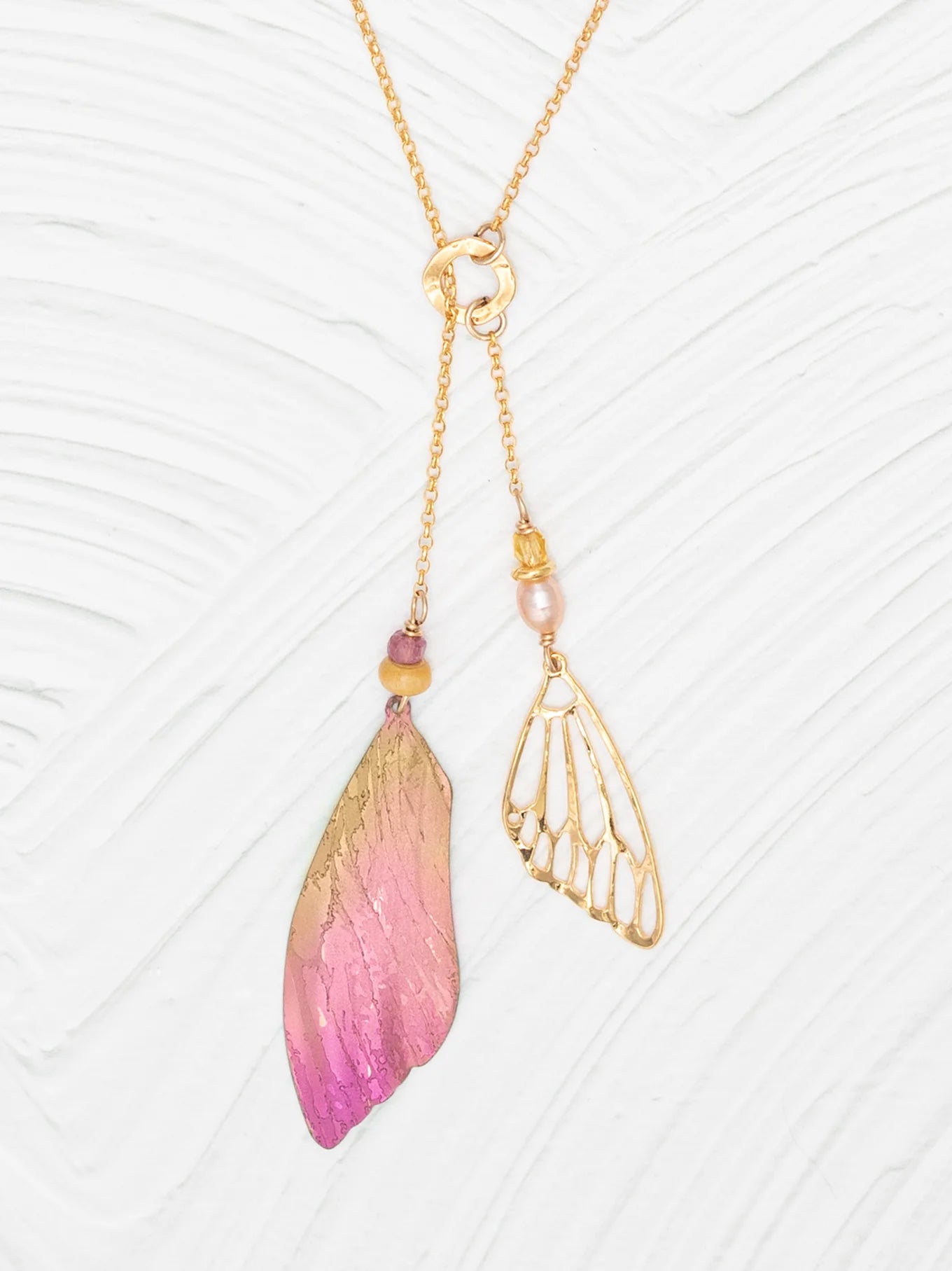 The Special Edition Flutterby Pink Lariat Necklace by Holly Yashi features an asymmetrical design: one side boasts a pink and gold ombré wing charm with beads, the other a delicate openwork gold wing with pearl, all set on an adjustable chain.