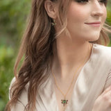 A woman with long brown hair smiles softly in a light cream dress, accessorized with turquoise dragonfly earrings by Holly Yashi. The blurred greenery behind her enhances her serene presence.