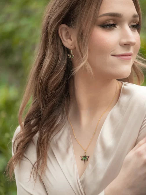 A woman with long brown hair smiles softly in a light cream dress, accessorized with turquoise dragonfly earrings by Holly Yashi. The blurred greenery behind her enhances her serene presence.