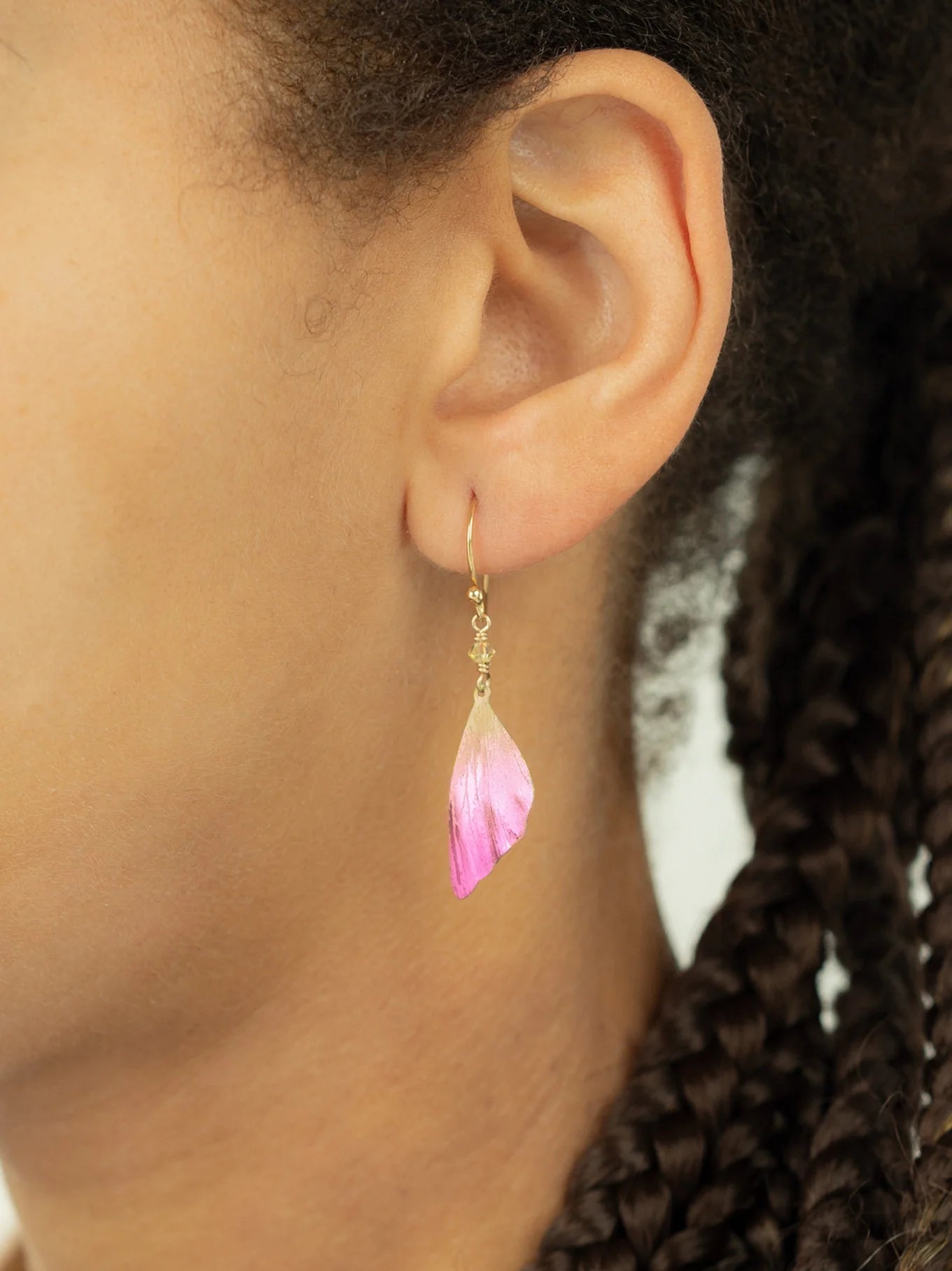 Close-up of a person’s ear wearing Special Edition Flutterby Pink Earrings by Holly Yashi, featuring a gold hook and pink and white petal-shaped charm. The earrings stand out beautifully against their braided hair, symbolizing cancer awareness.