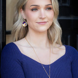 A woman with long blonde hair smiles softly in front of a dark background, wearing a blue ribbed top and Luna Earrings/Blue Moon by Holly Yashi—circular pendants featuring colorful abstract designs.