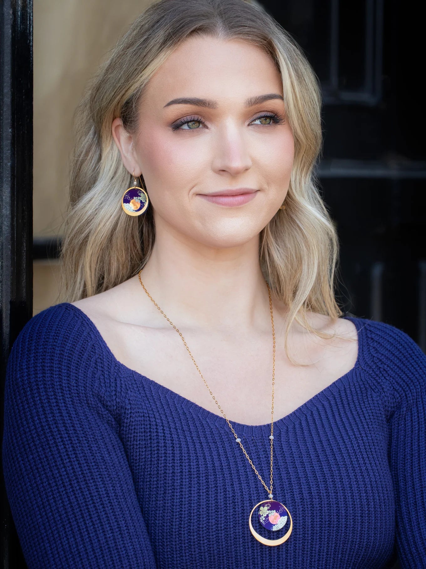 A woman with long blonde hair smiles softly in front of a dark background, wearing a blue ribbed top and Luna Earrings/Blue Moon by Holly Yashi—circular pendants featuring colorful abstract designs.
