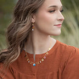 A woman with wavy brown hair wears a rust-colored blouse and matching earrings. Her look is enhanced by the Coral Reef Necklace from Holly Yashi Jewelry, adorned with various charms, as she gazes to the side against a blurred natural background.