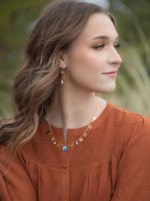 A woman with wavy brown hair wears a rust-colored blouse and matching earrings. Her look is enhanced by the Coral Reef Necklace from Holly Yashi Jewelry, adorned with various charms, as she gazes to the side against a blurred natural background.