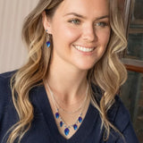 A smiling individual with wavy hair dons a navy sweater and layered necklaces featuring blue feather pendants. They complement the look with hypo-allergenic Healing Leaf Navy Earrings from Holly Yashi Jewelry while standing indoors near a wooden frame.