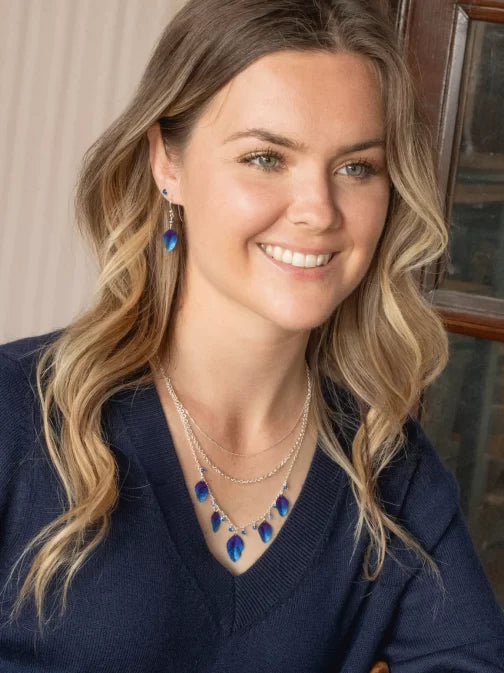 A smiling individual with wavy hair dons a navy sweater and layered necklaces featuring blue feather pendants. They complement the look with hypo-allergenic Healing Leaf Navy Earrings from Holly Yashi Jewelry while standing indoors near a wooden frame.