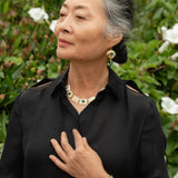 An older woman with gray hair in a bun stands outdoors before green shrubs with white flowers, wearing a black blouse and Holly Yashi's Estelle Emerald Earrings along with gold jewelry—a necklace and bracelet.