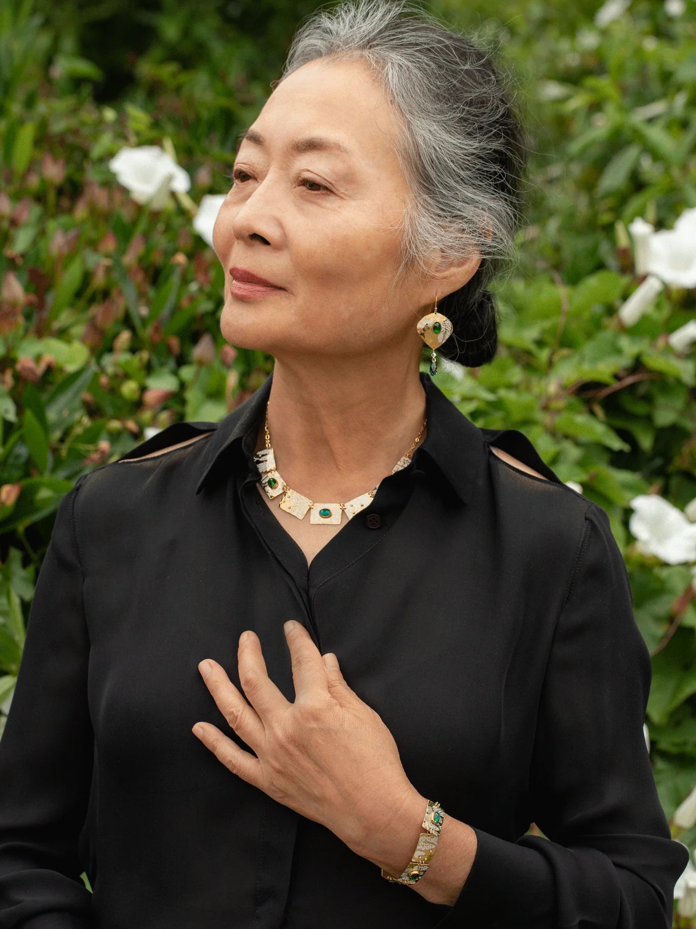 An older woman with gray hair in a bun stands outdoors before green shrubs with white flowers, wearing a black blouse and Holly Yashi's Estelle Emerald Earrings along with gold jewelry—a necklace and bracelet.