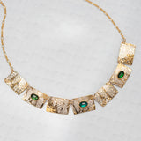 The Gilded Petra Emerald Necklace by Holly Yashi showcases seven hammered rectangles, three with emerald green crystals, on a delicate gold chain. This Victorian-inspired piece shines with elegance on a light textured backdrop.