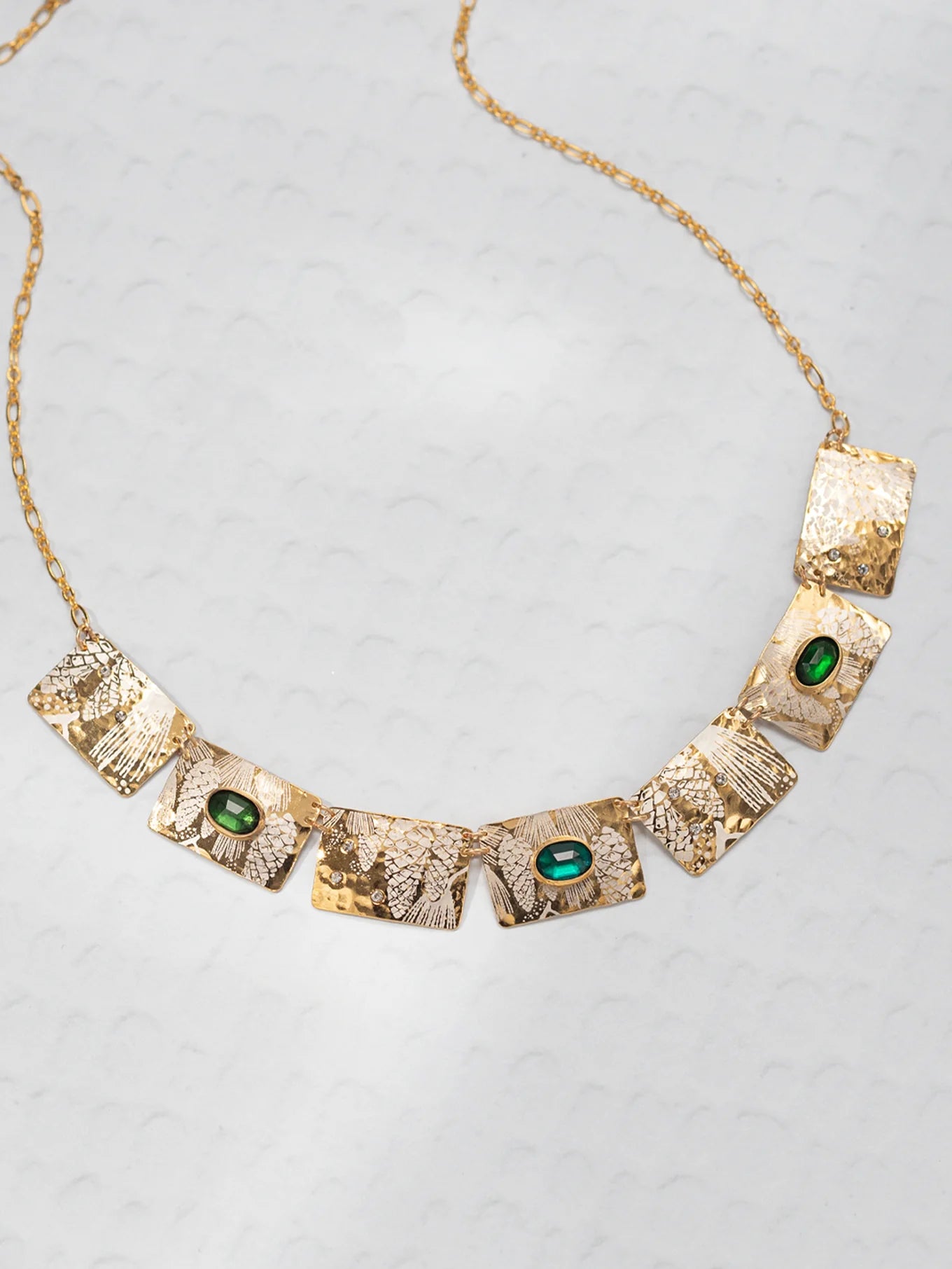 The Gilded Petra Emerald Necklace by Holly Yashi showcases seven hammered rectangles, three with emerald green crystals, on a delicate gold chain. This Victorian-inspired piece shines with elegance on a light textured backdrop.
