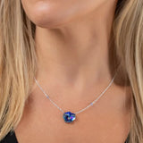 A person, wearing a black top, elegantly wears a Bonnie Blue Garden Pansy Necklace from Holly Yashi. Their long blonde hair cascades over the shoulders, highlighting the shimmering silver chain and pendant. This stylish hypo-allergenic piece is both exquisite and comfortable.
