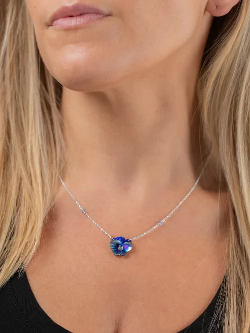 A person, wearing a black top, elegantly wears a Bonnie Blue Garden Pansy Necklace from Holly Yashi. Their long blonde hair cascades over the shoulders, highlighting the shimmering silver chain and pendant. This stylish hypo-allergenic piece is both exquisite and comfortable.