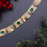 The Gilded Petra Emerald Bracelet by Holly Yashi, featuring gold square links and emerald green crystals, is displayed on a dark surface with red berries and frosted greenery for a festive touch.