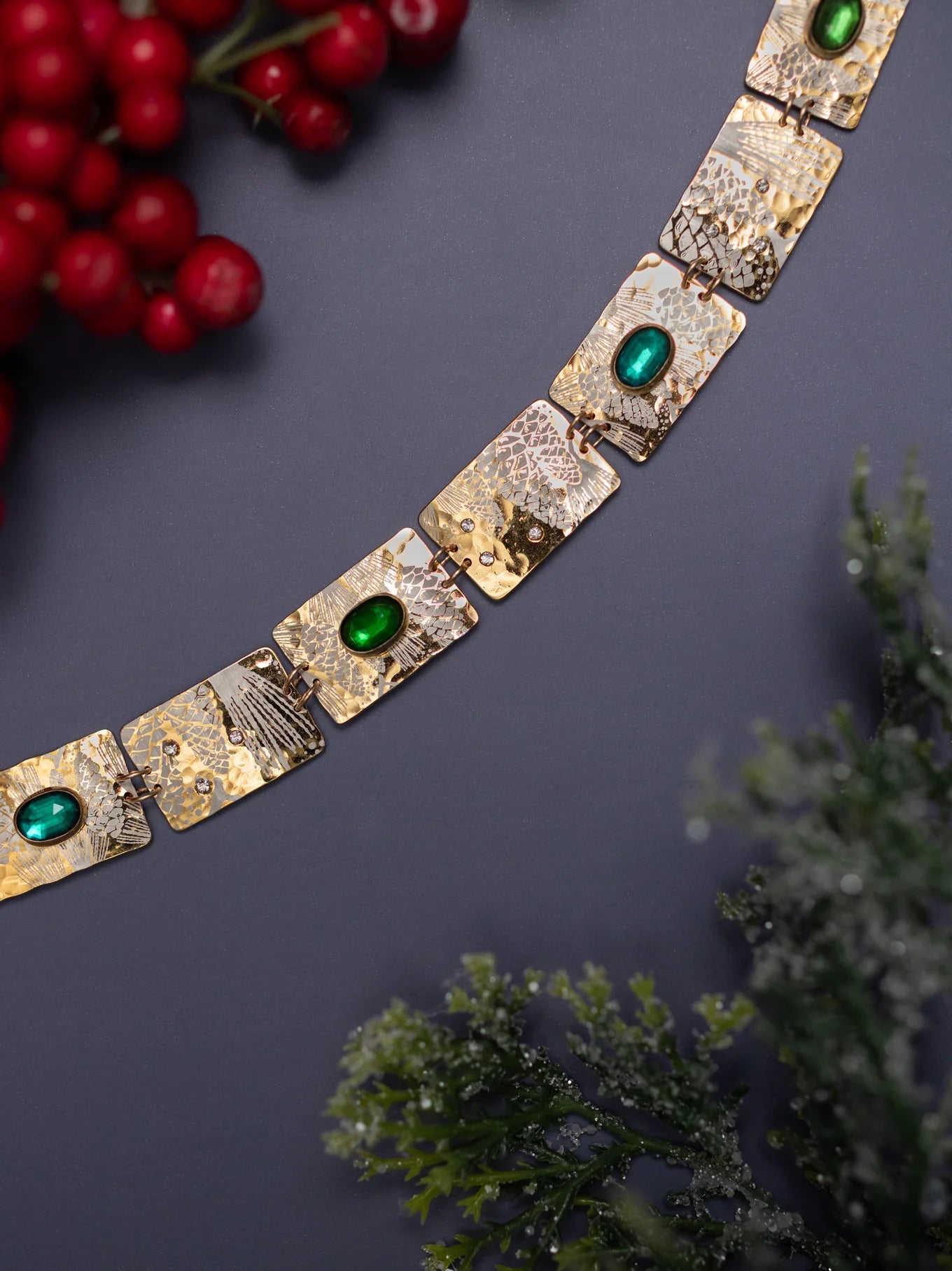 The Gilded Petra Emerald Bracelet by Holly Yashi, featuring gold square links and emerald green crystals, is displayed on a dark surface with red berries and frosted greenery for a festive touch.