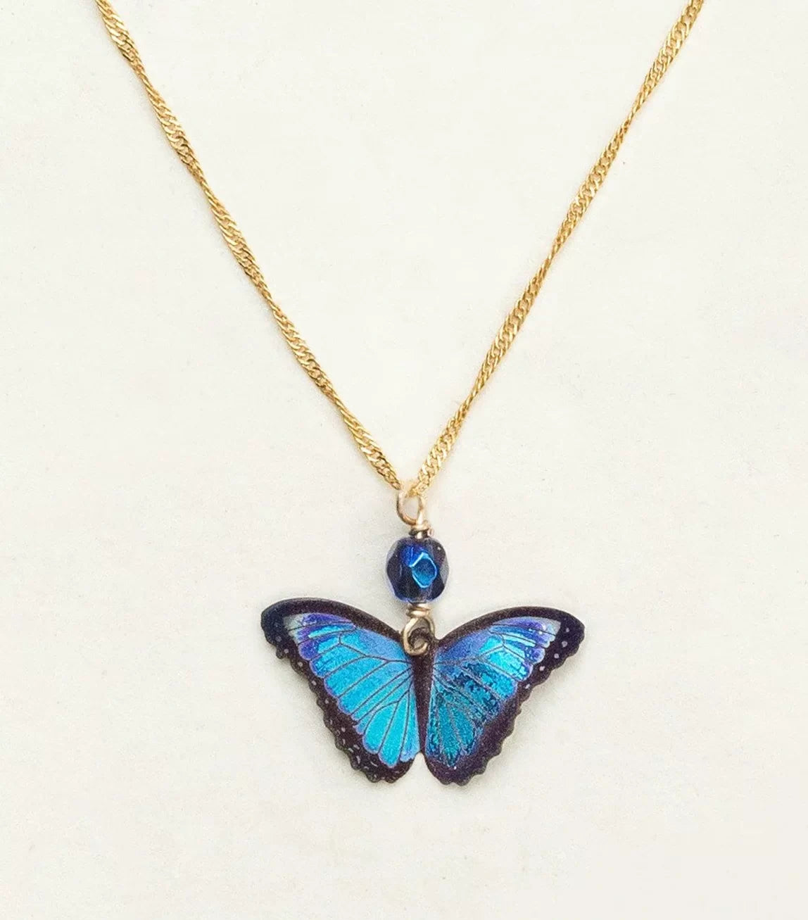 Introducing the Blue Bella Butterfly Necklace by Holly Yashi: a gold-plated chain featuring an iridescent blue butterfly pendant with a small dark blue bead above it, set against a light background for timeless allure.