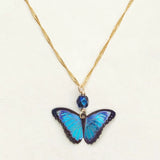 Introducing the Blue Bella Butterfly Necklace by Holly Yashi: a gold-plated chain featuring an iridescent blue butterfly pendant with a small dark blue bead above it, set against a light background for timeless allure.