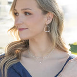 A woman with long, wavy blonde hair glances to the side while outdoors. She wears a blue top and Holly Yashis Rosa Gold and Pearls Hoop Earrings, which glint in the light, paired with layered necklaces of freeform pearls. Bracelets jingle on her wrist against a sunny, softly blurred background.
