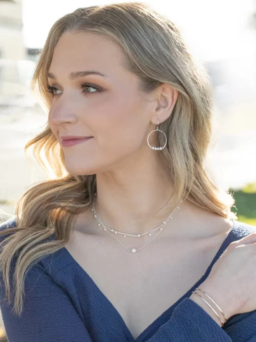 A woman with long, wavy blonde hair glances to the side while outdoors. She wears a blue top and Holly Yashis Rosa Gold and Pearls Hoop Earrings, which glint in the light, paired with layered necklaces of freeform pearls. Bracelets jingle on her wrist against a sunny, softly blurred background.