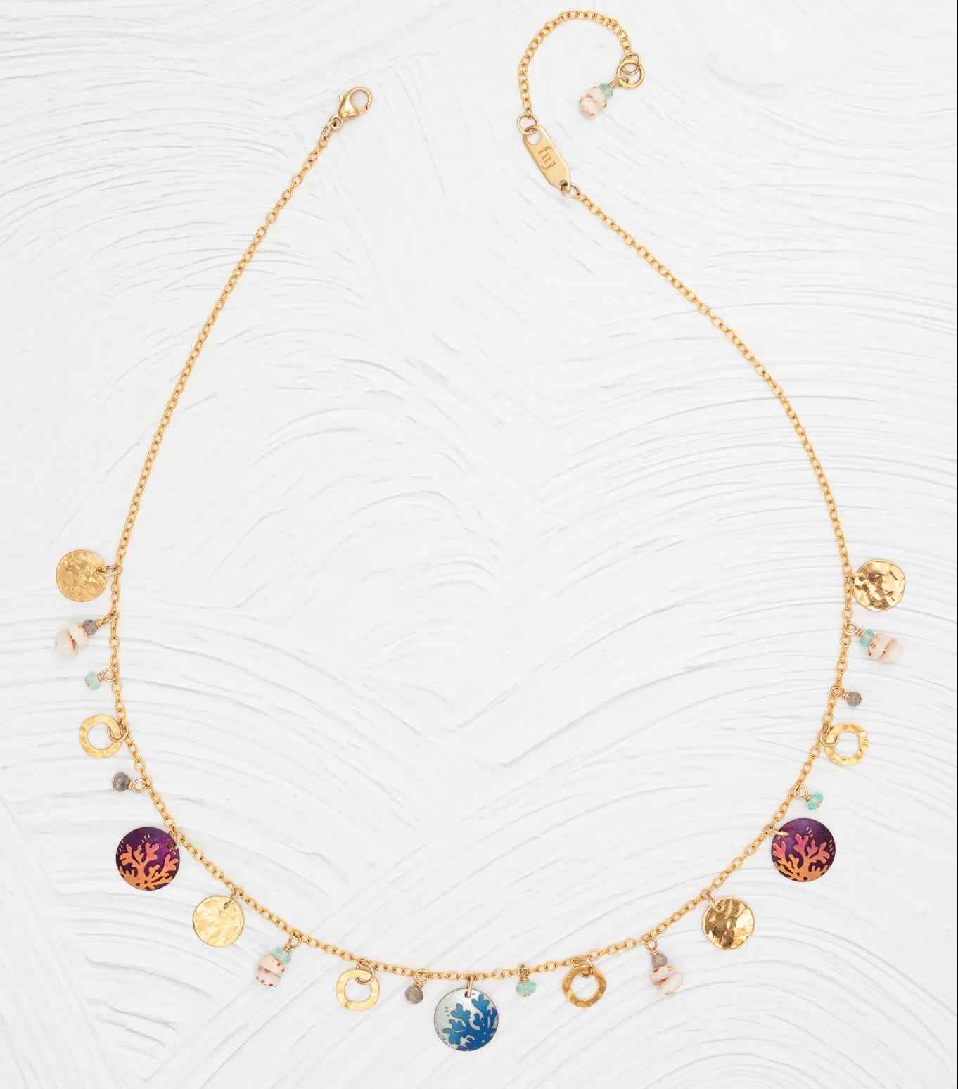 The Holly Yashi Coral Reef Necklace boasts a gold-plated chain with colorful circle pendants, small gold discs, and pastel beads arranged on a white textured surface.