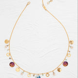 The Holly Yashi Coral Reef Necklace boasts a gold-plated chain with colorful circle pendants, small gold discs, and pastel beads arranged on a white textured surface.